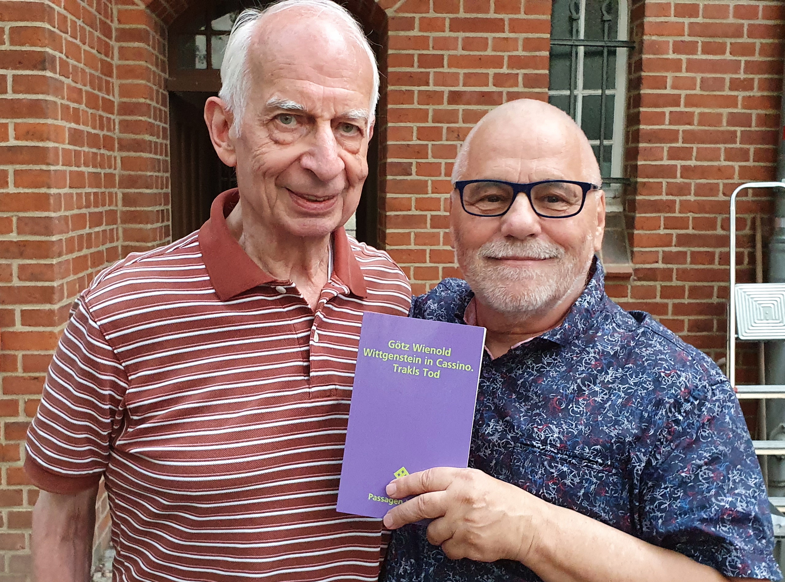 Zwei Männer posieren mit einem lila Buch vor einer Ziegelmauer.
